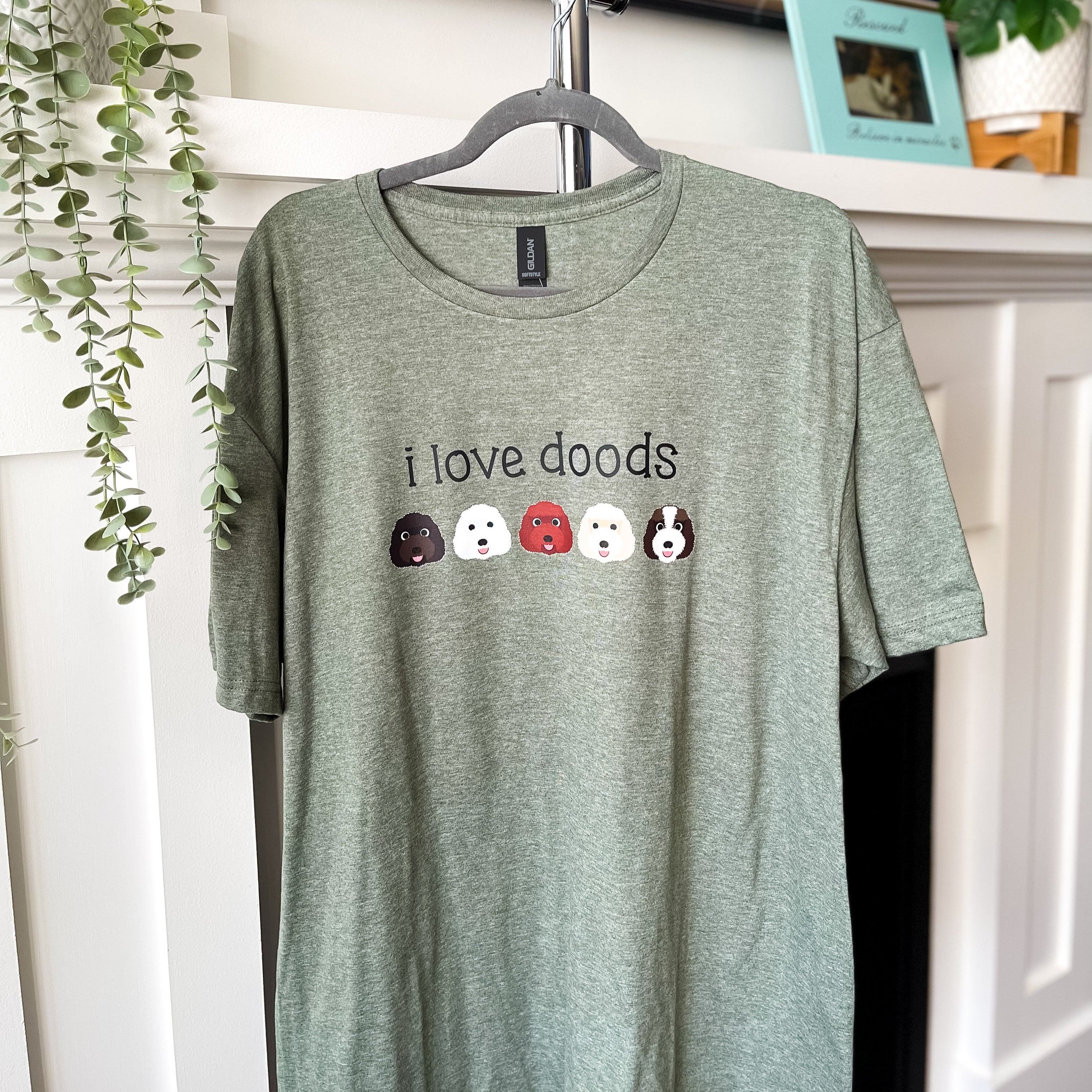 Green t-shirt with 'i love doods' text and dog icons on a hanger against a home decor background.