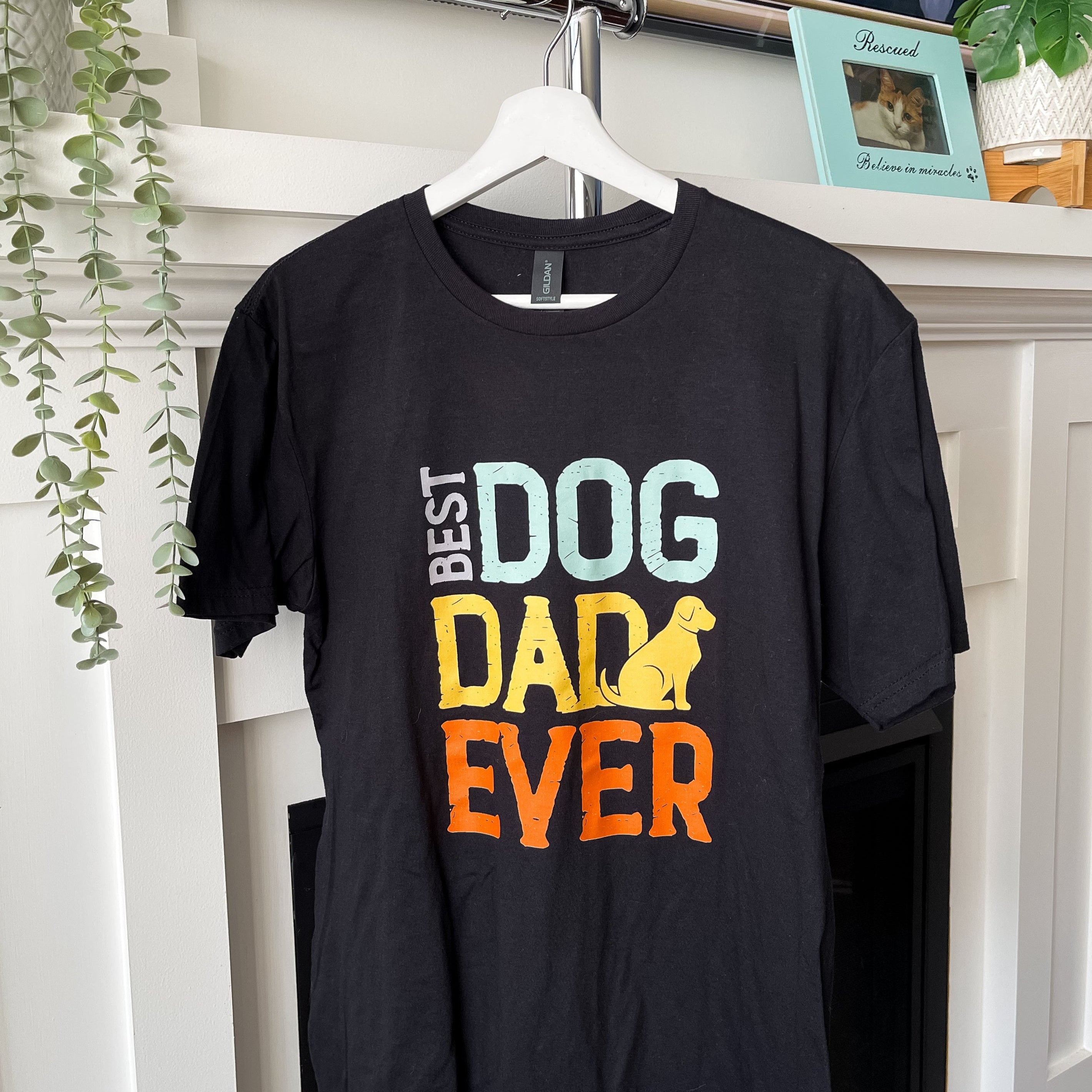 Black t-shirt with 'Best Dog Dad Ever' text on a hanger against a white wall.