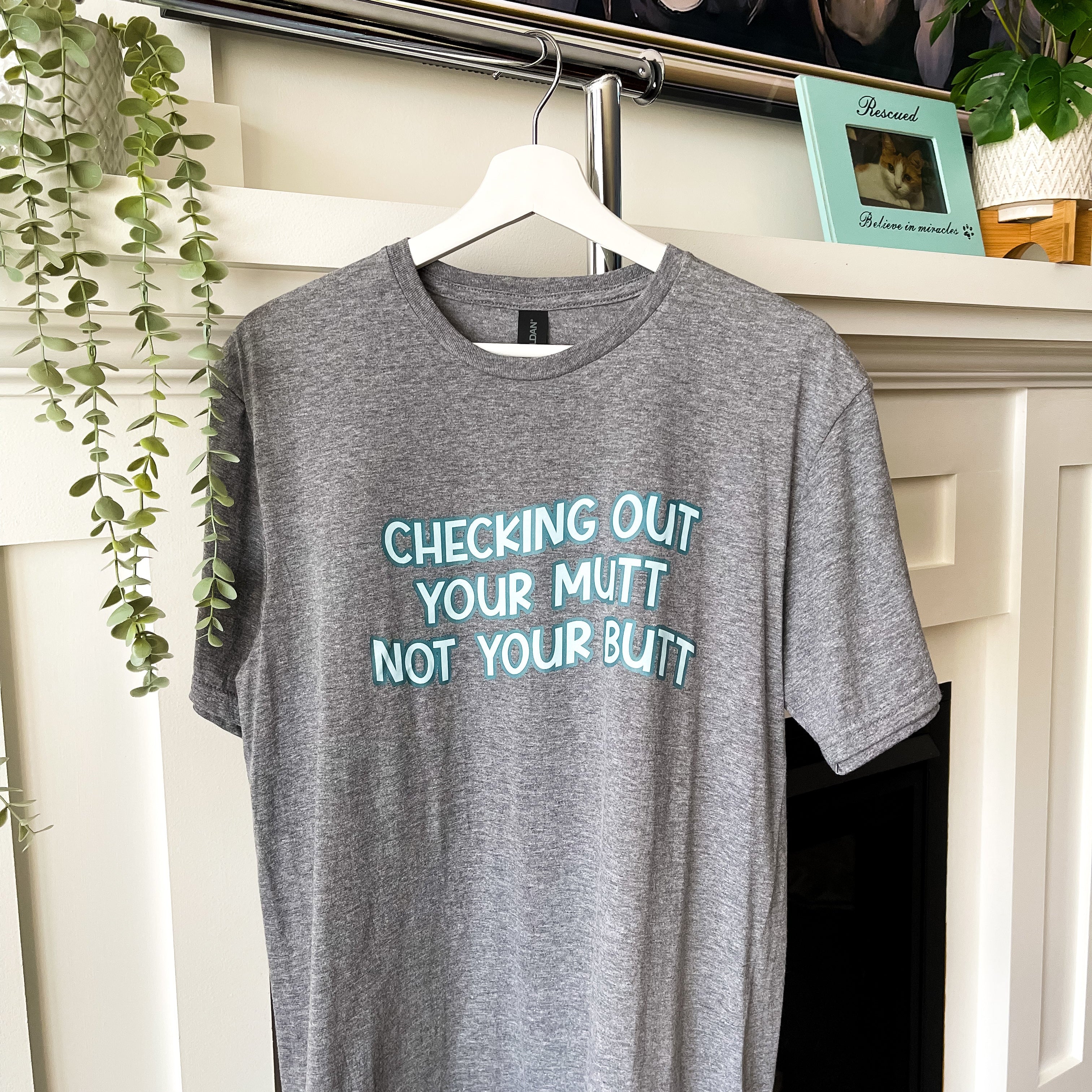Gray t-shirt with humorous text on a hanger in front of a fireplace.