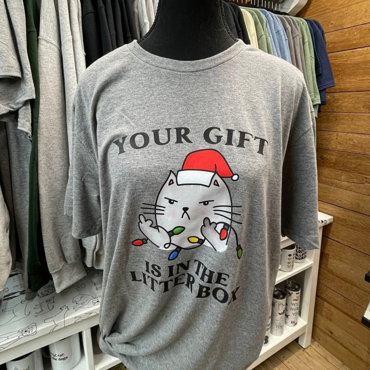 A gray t-shirt with a graphic print of a cat wearing a Santa hat and the text 'YOUR GIFT IS IN THE LITTER BOX'.