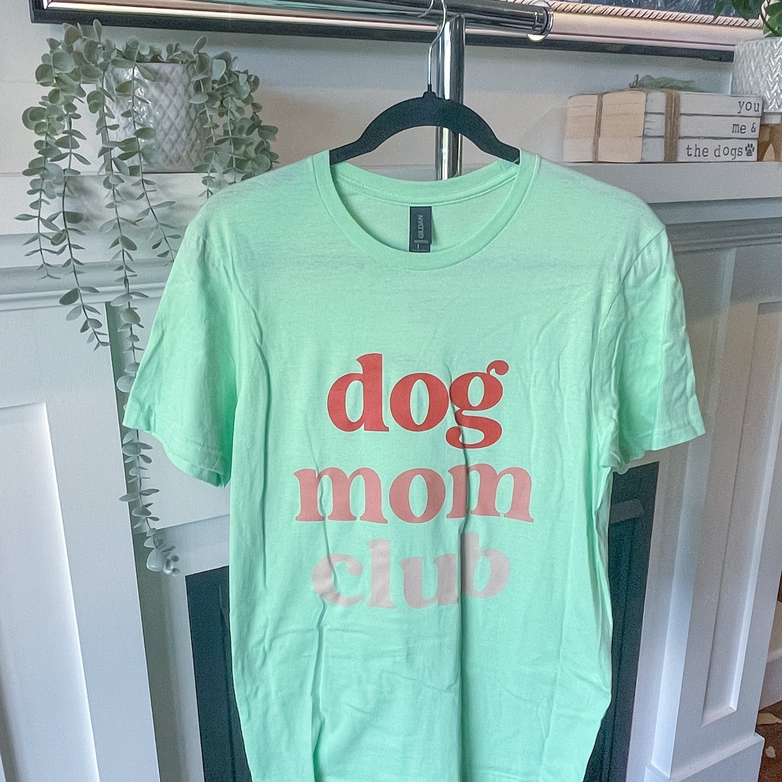 Light green t-shirt with 'dog mom club' text on a hanger in a room.
