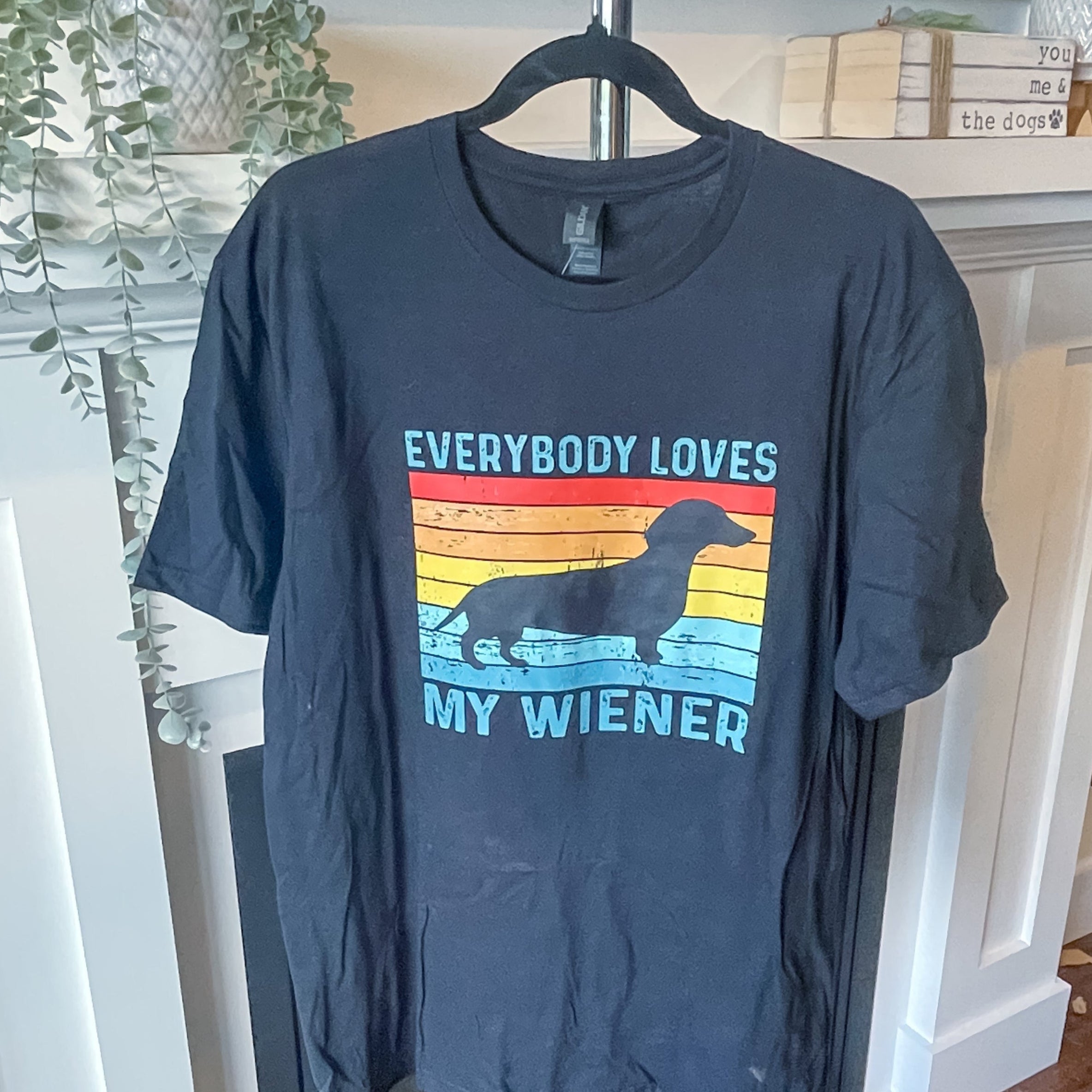 Black t-shirt with 'Everybody Loves My Wiener' graphic on a hanger in a room.