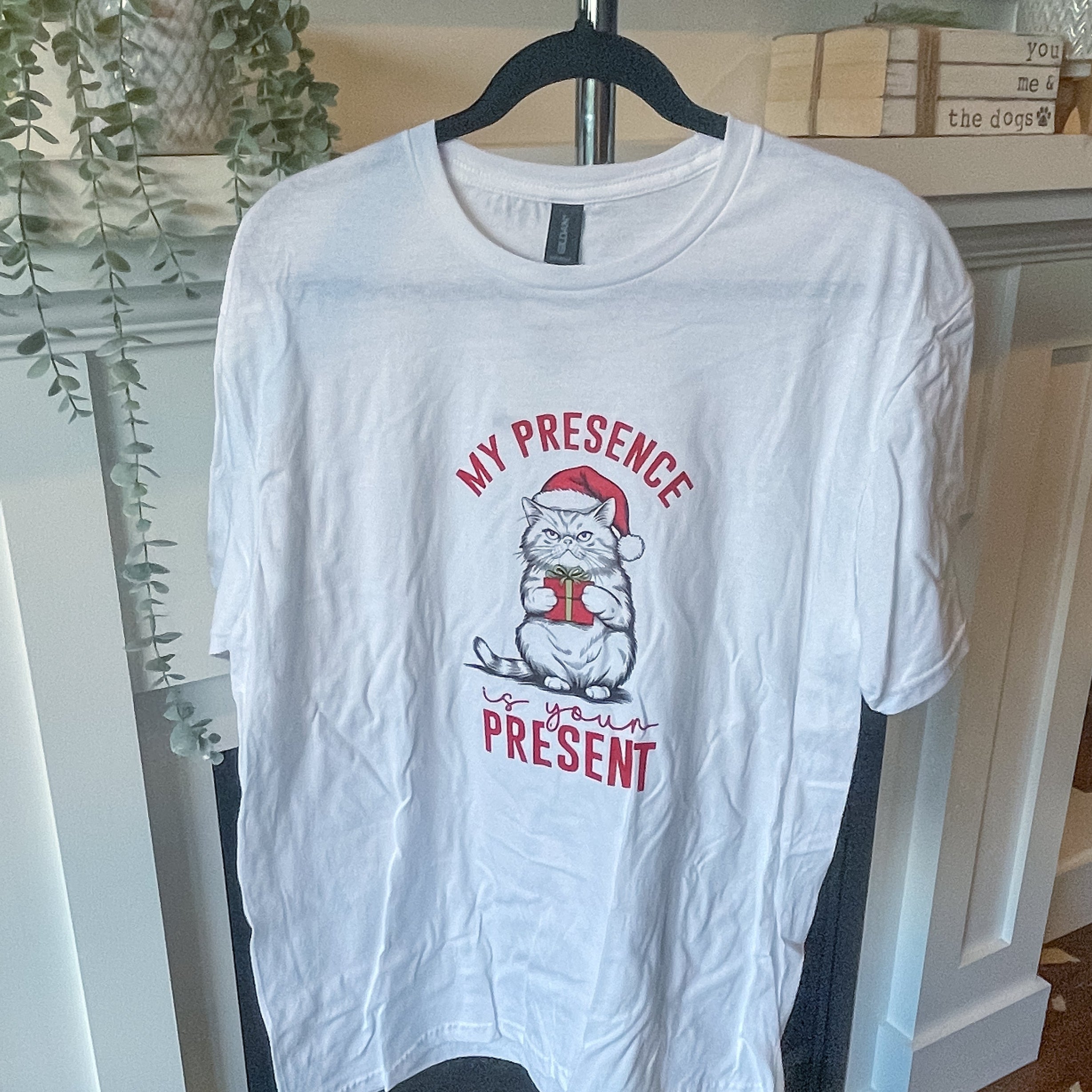 White t-shirt with red graphic of a grumpy cat holding a present and the test reads "My presence is your present" on a hanger against a decorative wall.